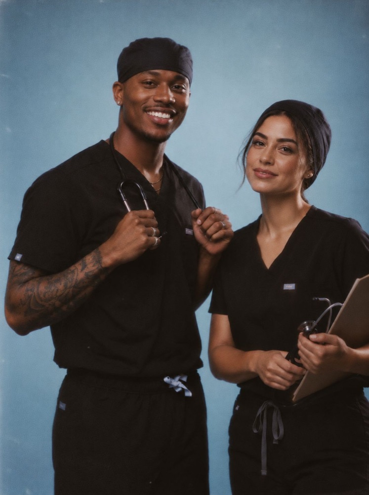 Médico e médica de pijama cirúrgico preto sorrindo; ela segura uma prancheta