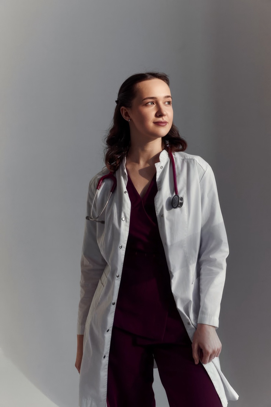 Médica de jaleco branco e estetoscópio vinho, olhando para o lado com um leve sorriso
