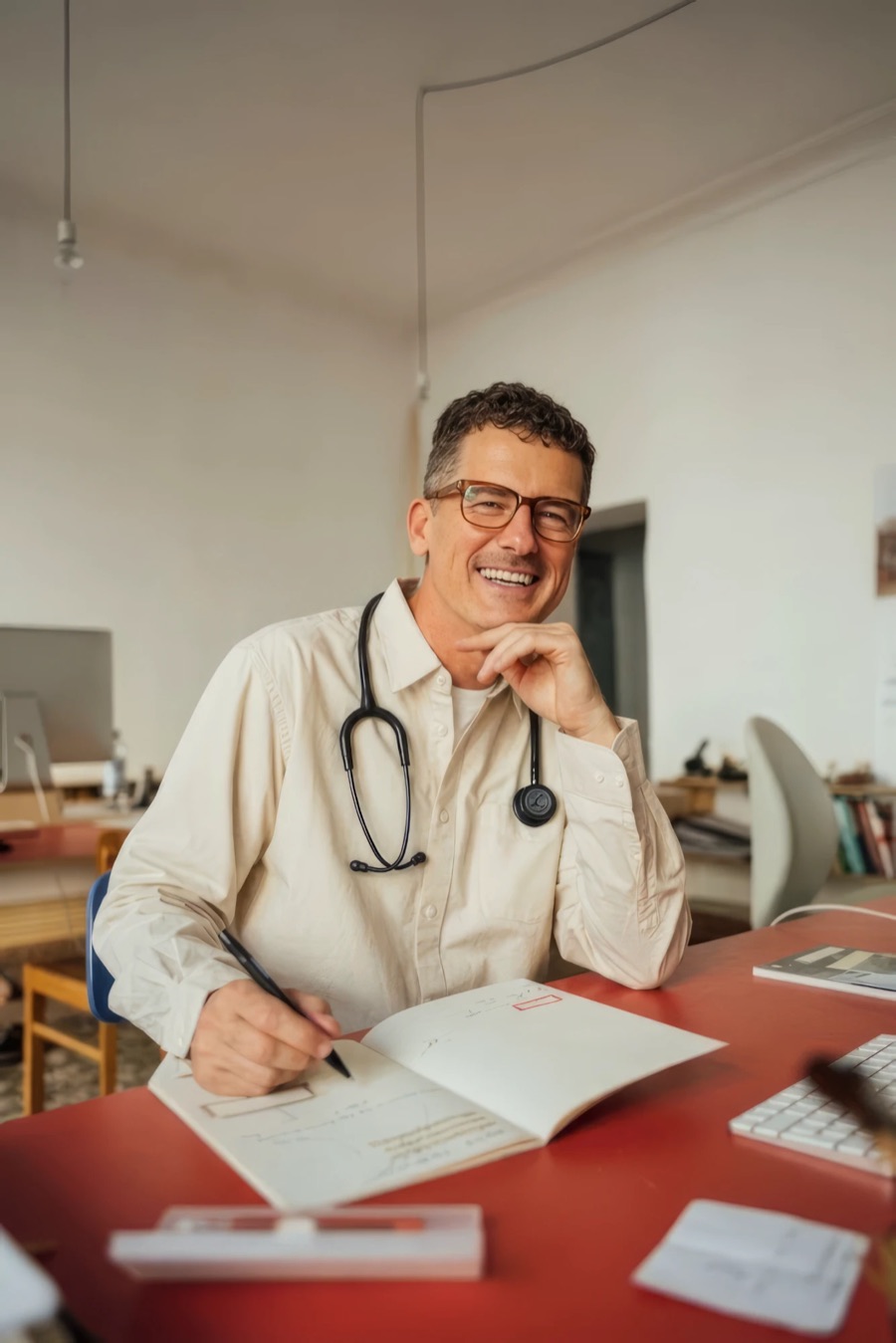 Médico de óculos e camisa clara, com estetoscópio no pescoço, sorrindo sentado à mesa enquanto faz anotações em um caderno